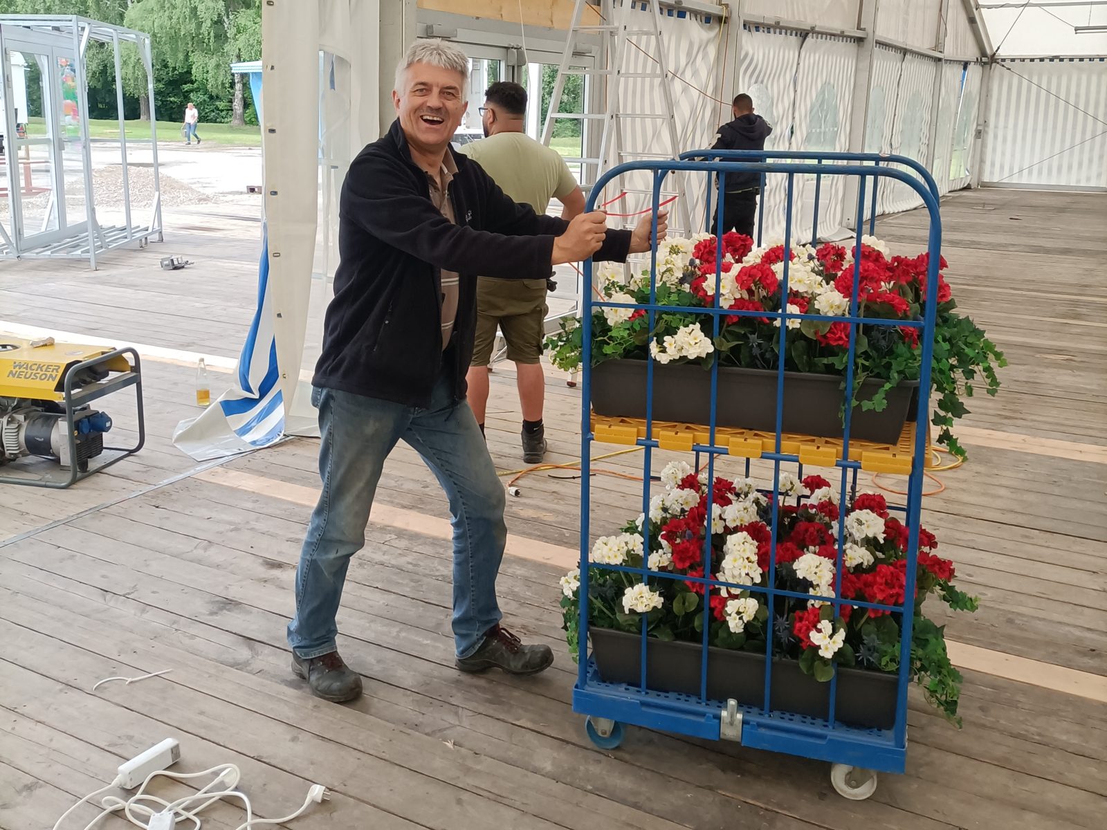 Sepp schiebt beim Aufbau im Festzelt einen Rollwagen mit Blumenkästen